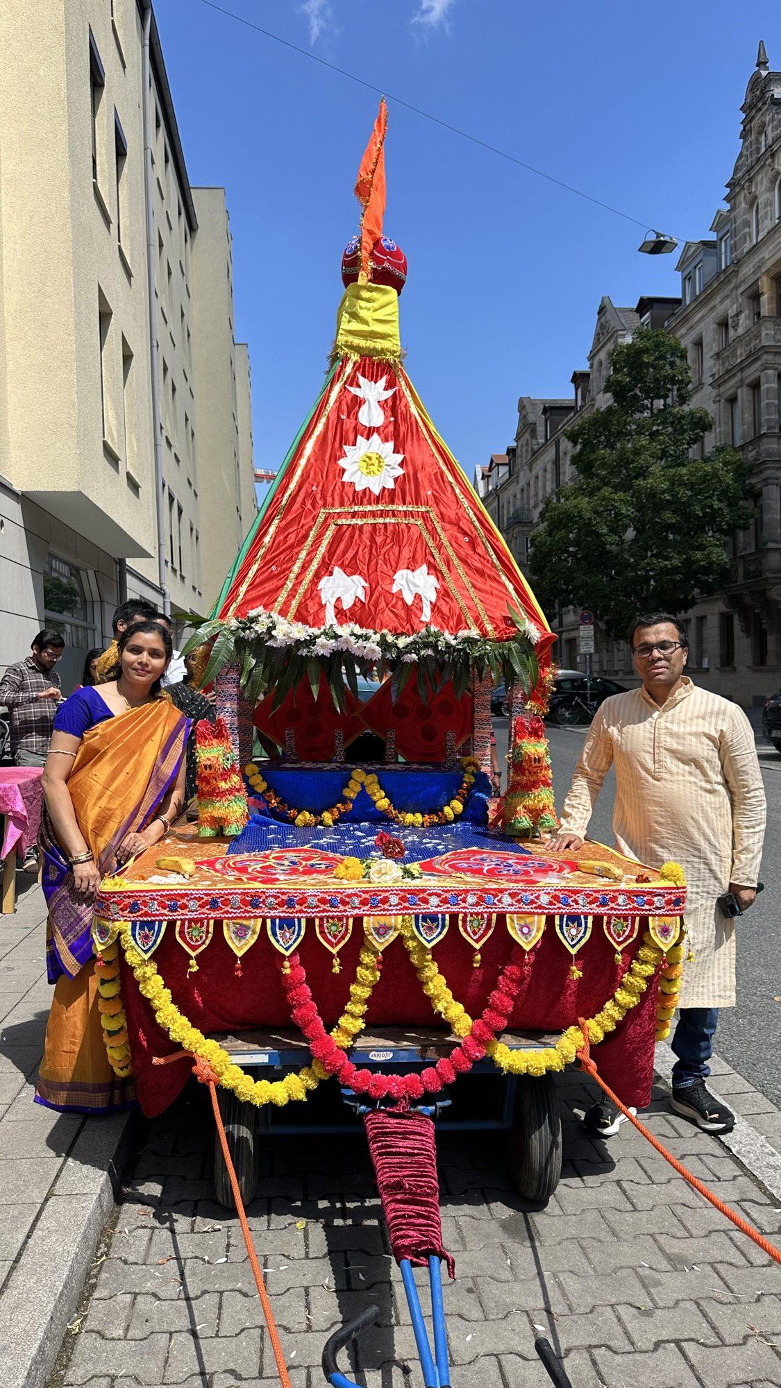 Two community members standing beside a small decorated Rath Yatra chariot with flower garlands on a city street